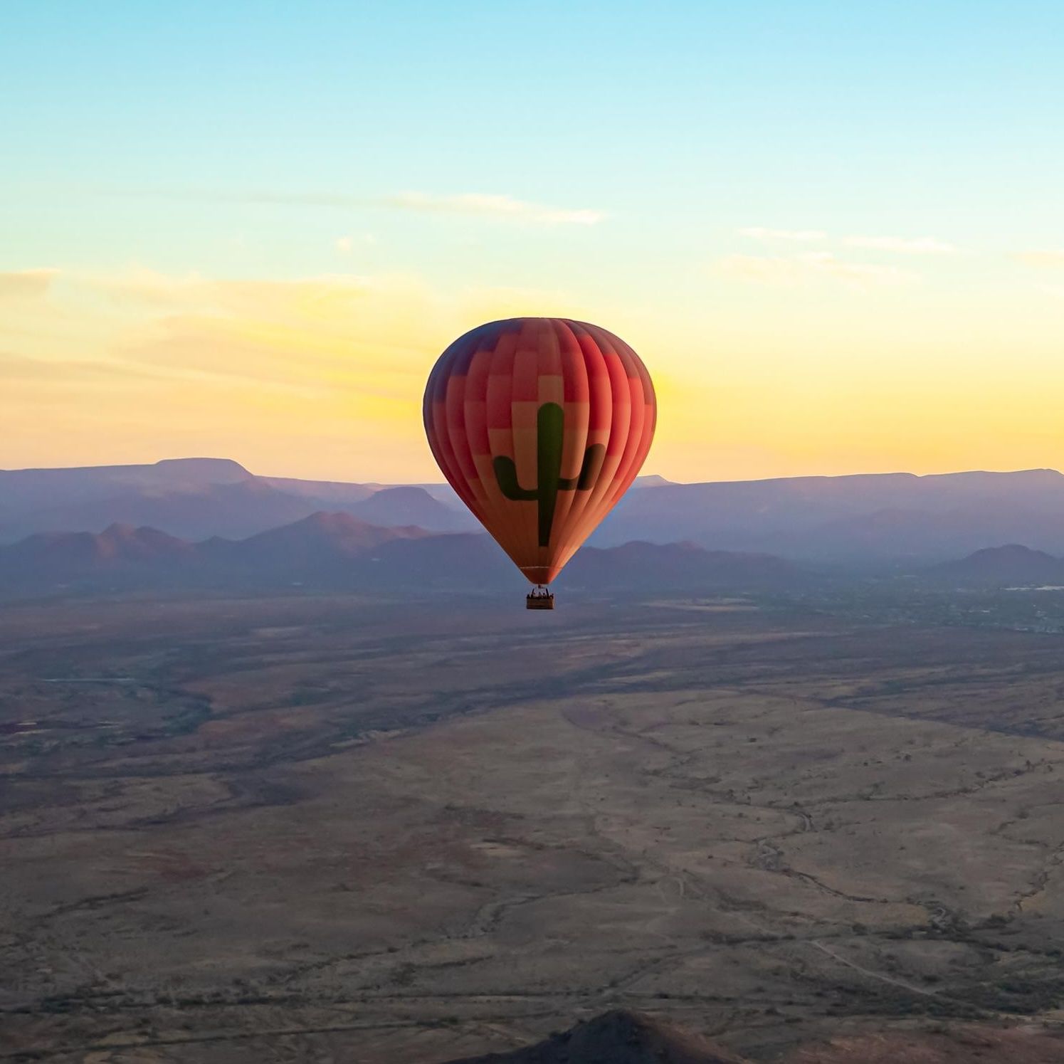 A hot air balloon with a cactus design floats over a desert landscape at sunrise or sunset.