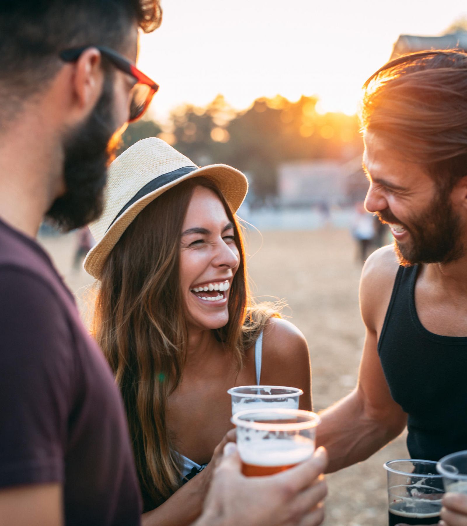 Four friends laugh and hold drinks outdoors at sunset, enjoying a casual social gathering.