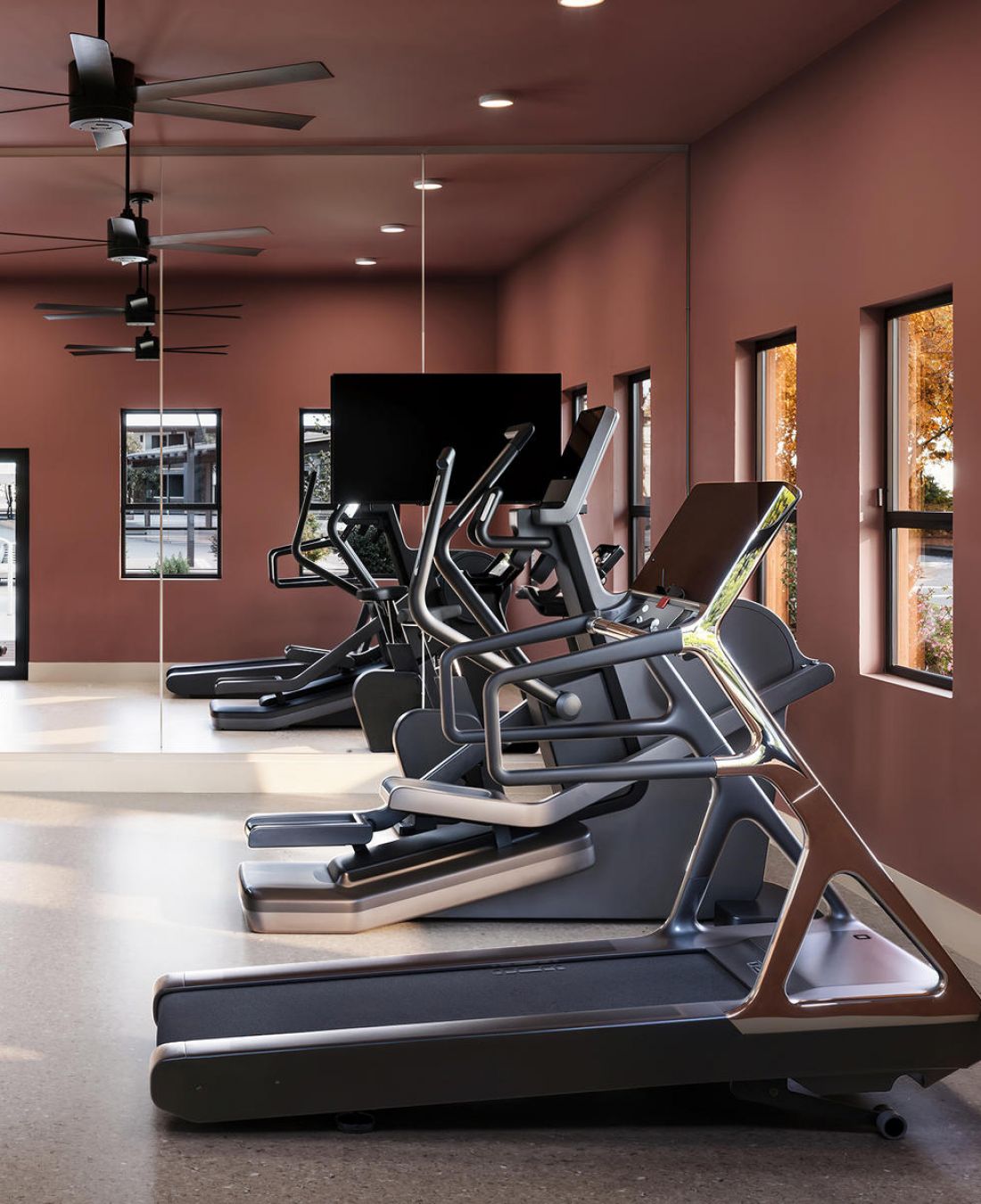 Modern gym with treadmills and ellipticals facing mirrored wall and windows with autumn trees outside.