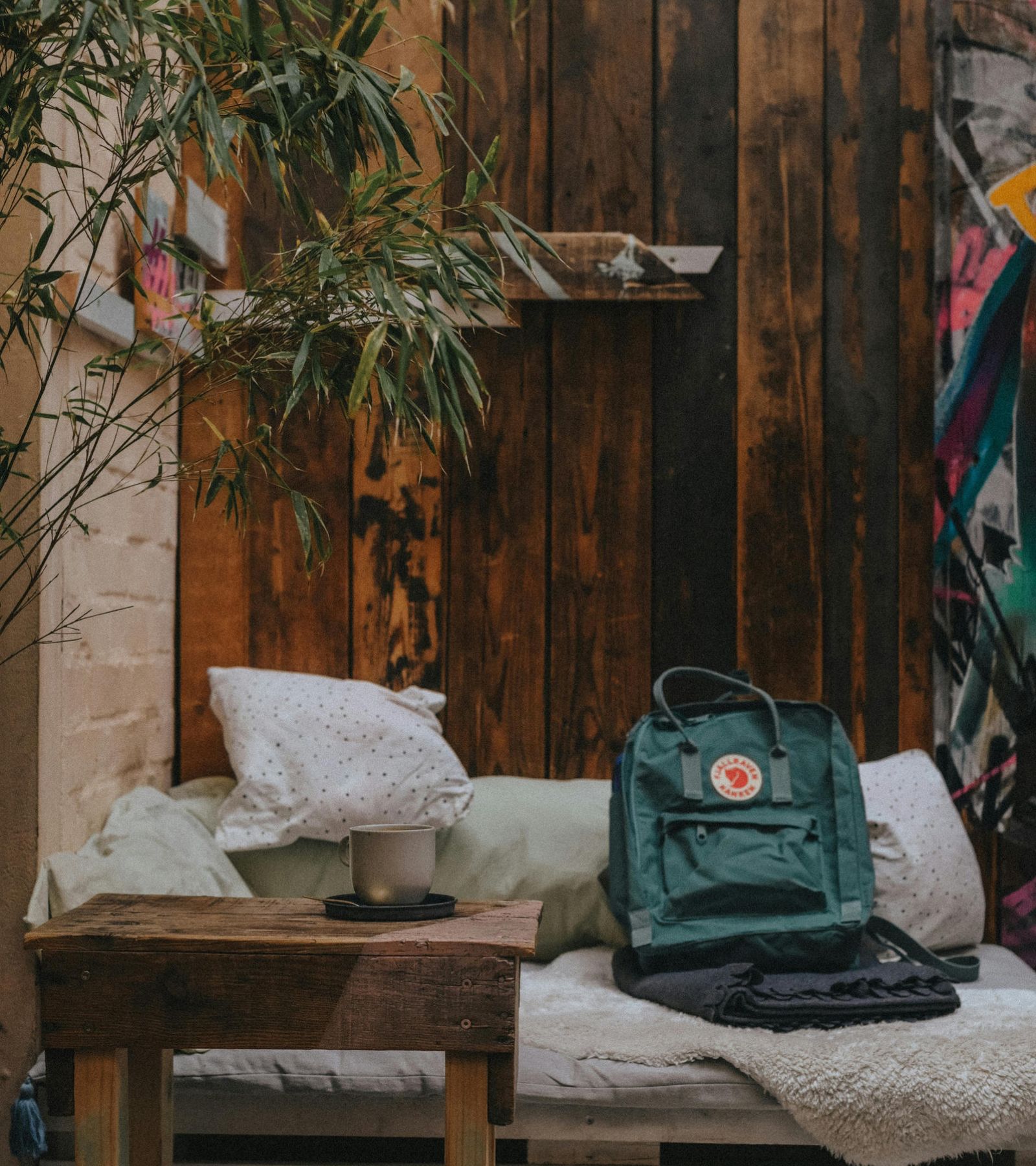Cozy nook with pillows, a green backpack, and coffee cup on a wooden table next to rustic wooden walls.
