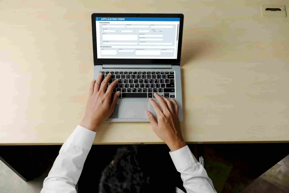 Person filling out an online application form on a laptop at a desk, viewed from above.