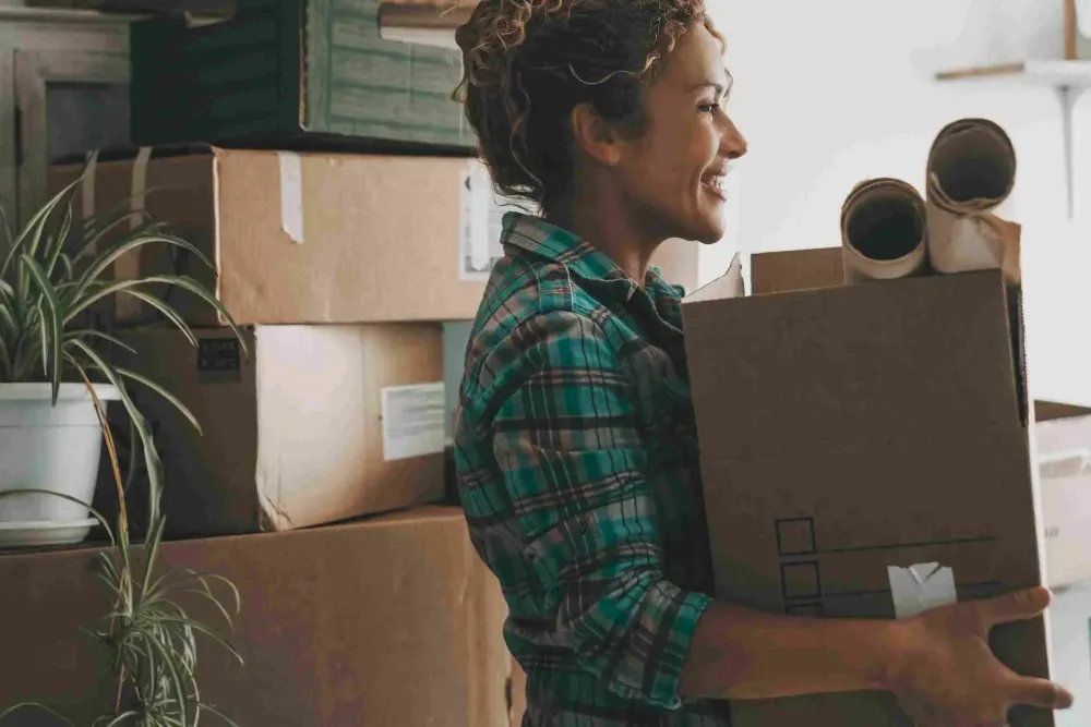 Smiling woman holding a moving box stands near stacked cardboard boxes and rolled-up papers.