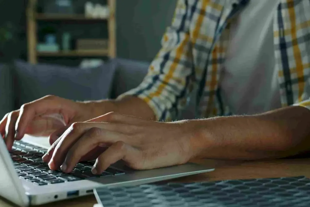 Person typing on a laptop keyboard, wearing a plaid shirt, with a blurred background.