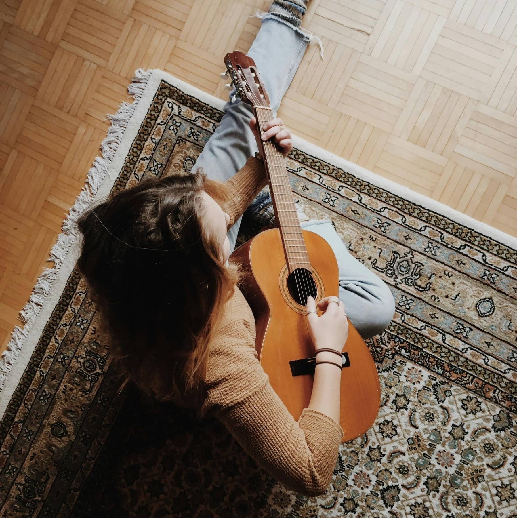 Person sitting on a patterned rug, playing an acoustic guitar, viewed from above.